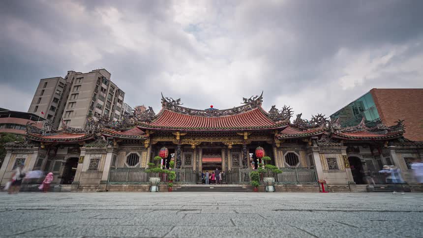 A wide view of a traditional temple with an ornate roof as clouds move across a grey sky and people walk through a paved courtyard with blurred motion