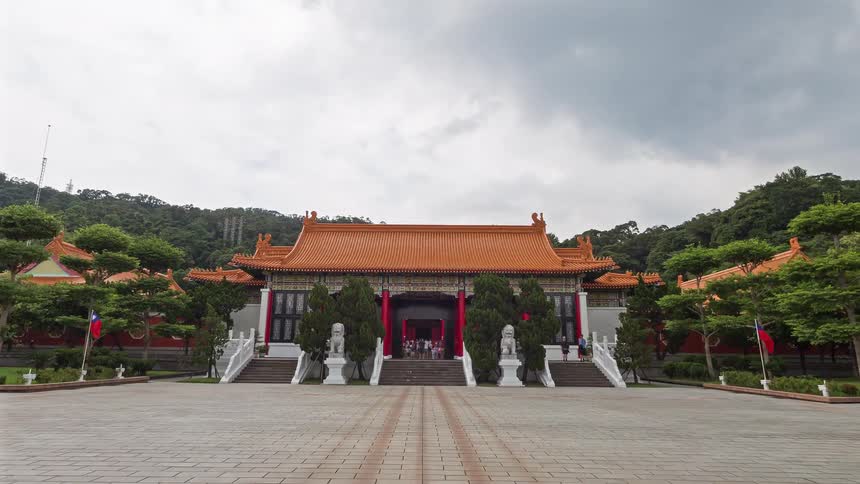 A camera moves quickly forward across a large stone courtyard toward a traditional temple with an orange roof and red pillars under a cloudy sky while tourists walk through the open gates and around the historic grounds