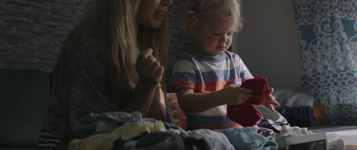 A toddler boy unfolding clean clothes while sitting next to his mother indoors.