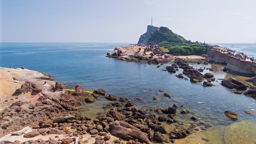 A rocky shoreline with unique sandstone formations leading toward a lush green hill under a clear sky while tourists walk along a paved path near the calm blue ocean water during a bright day at a coastal park