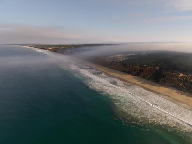 Ocean waves reaching a sandy shoreline along vegetationcovered cliffs with low fog over the coast
