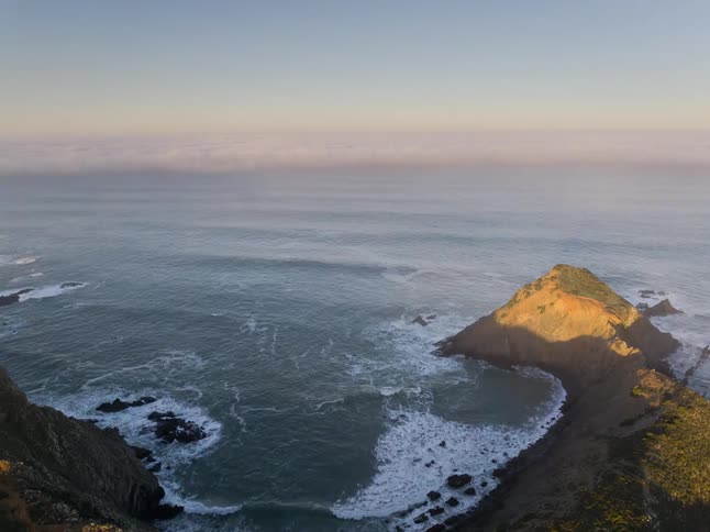 Golden sunlight hitting a steep coastal cliff as white waves crash against dark rocks along a rugged shoreline under a clear morning sky with a thick layer of low clouds resting on the distant ocean horizon