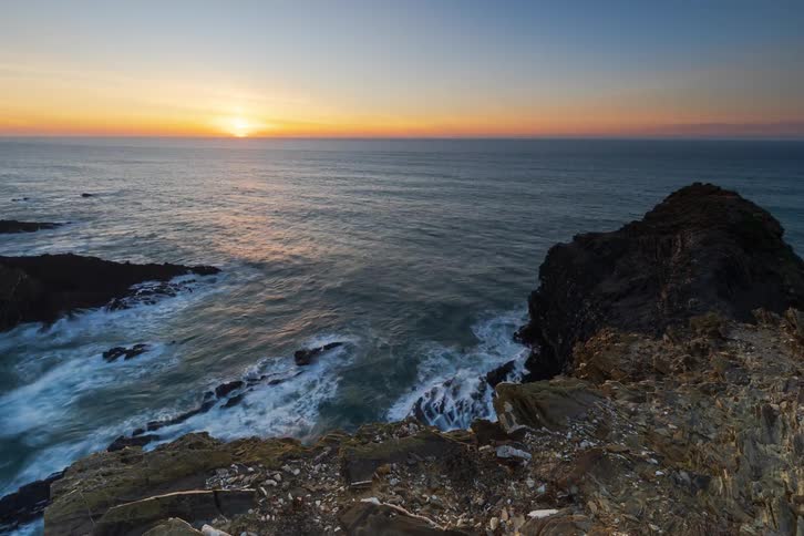 Sun on the horizon over the ocean with waves hitting rocks and a dark cliff in the foreground