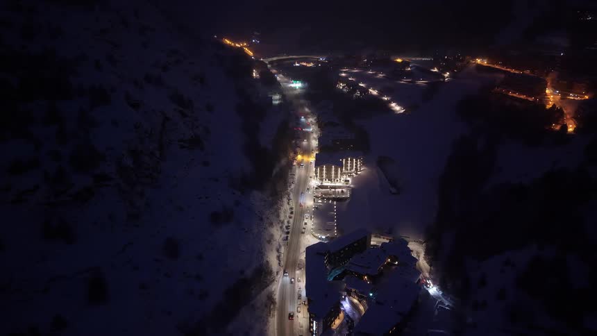 Illuminated road crossing a snowy valley with buildings and vehicles at night