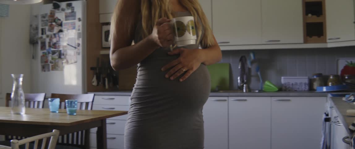 Expectant mother carrying a warm drink while walking in a bright and airy kitchen.