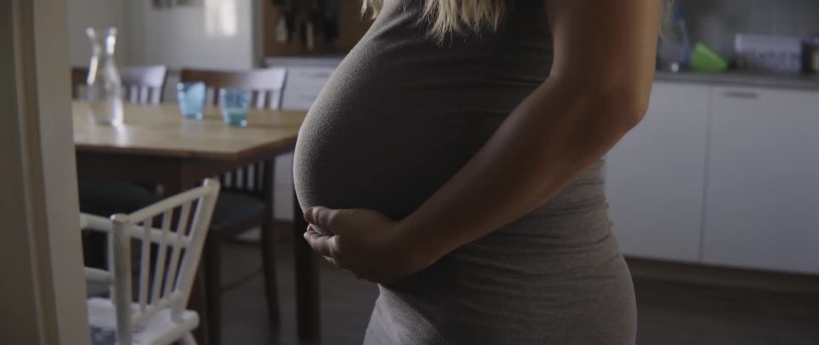 Young pregnant woman standing in a sunny kitchen while touching her baby bump tenderly.