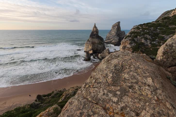People walk near rocks along the coast as waves roll into shore and over the beach