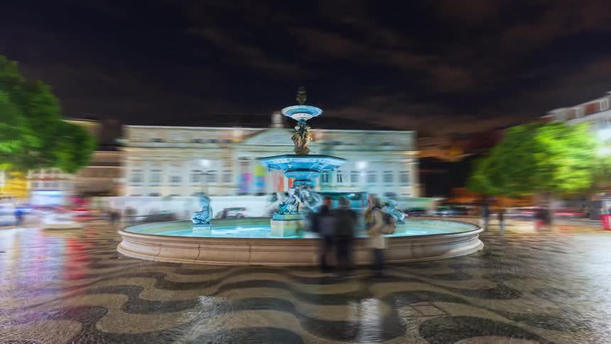 People move through a brightly lit plaza containing a water fountain at night