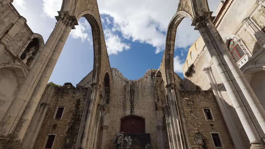 A roofless towering gothic stone arches and weathered walls against a bright blue sky filled with clouds while a few visitors walk across the stone floor below