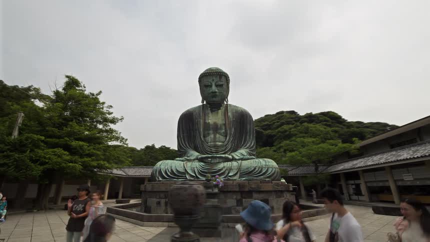 A large bronze statue sits in the center of a stone courtyard at a temple under a cloudy sky while tourists walk around the base and green trees cover the surrounding hills