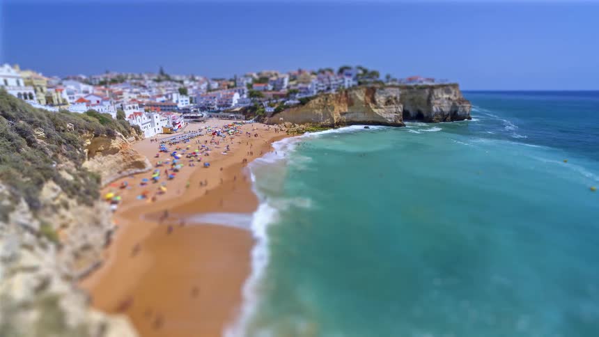 A crowded sandy beach nestled between a white coastal village and steep tan cliffs as turquoise ocean waves crash against the shore under a clear blue sky during a bright summer day