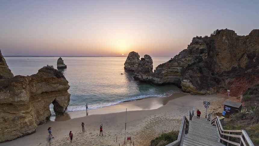 The sun rising over a calm ocean bay framed by massive limestone cliffs and sea stacks while a few people gather on the sand near a wooden staircase and a small building at the edge of the beach