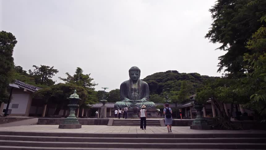 A large bronze statue of the great buddha of kamakura sits in a courtyard surrounded by trees and buildings while people walk nearby on a cloudy day in japan