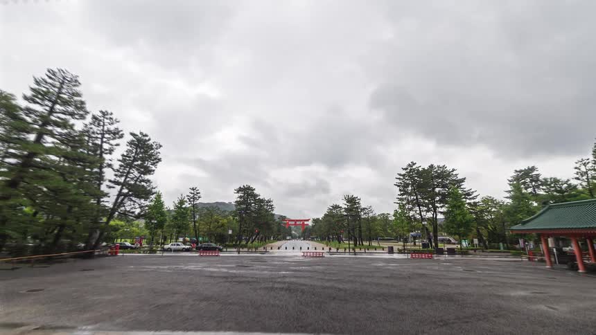 A wide time lapse shot moves backward through a massive orange gate to reveal the grand wooden facade of a japanese shrine under a grey overcast sky while people walk across the wet ground during a rainy day