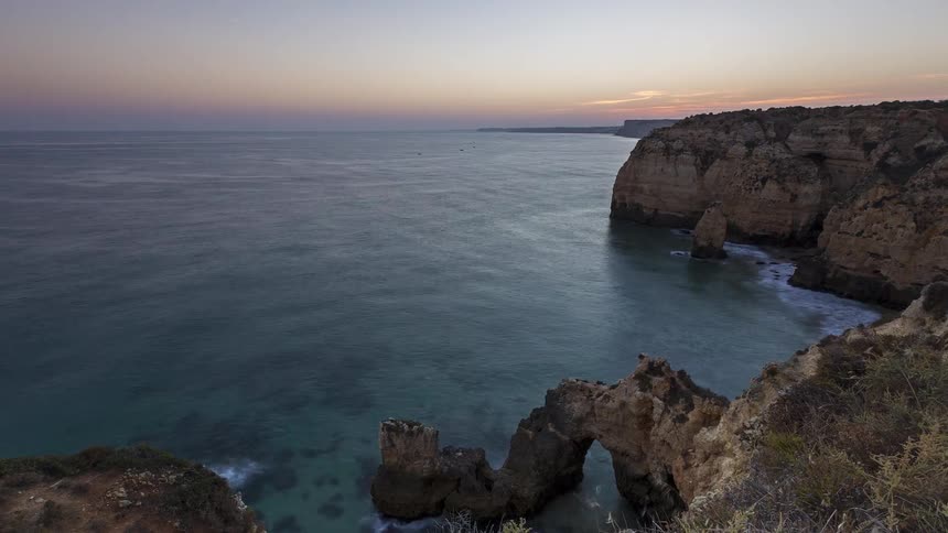 Mountain cliffs with an arch rock formation along the coast as waves roll into shore and boats sail on the water at sunset