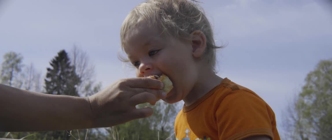 Mother and toddler boy sharing a snack together while sitting in a grassy field.