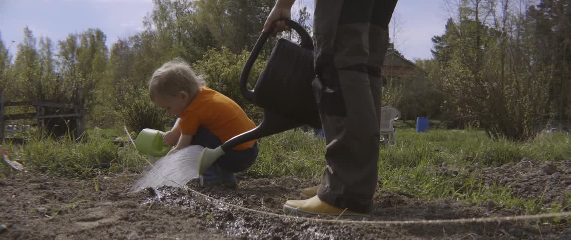 Woman and toddler boy pouring water from watering cans onto dirt in garden