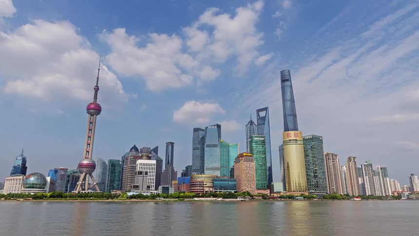 A wide view of the shanghai skyline along the huangpu river featuring the oriental pearl tower and shanghai tower under a blue sky with moving clouds as boats pass by the waterfront
