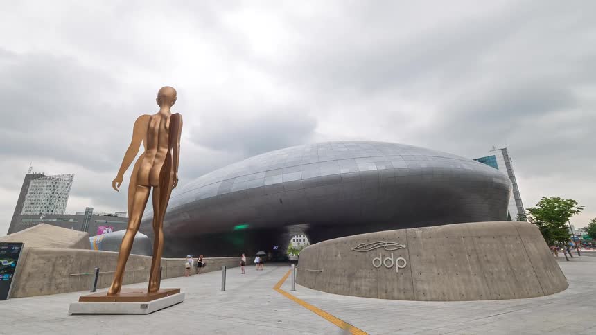 Golden statue in a plaza in front of a futuristic ovalshaped building under a cloudy sky