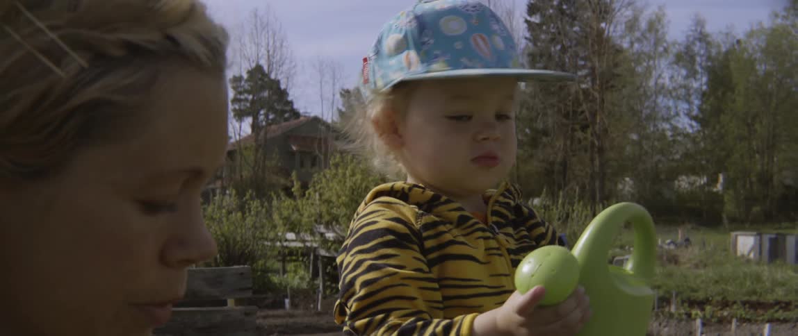Toddler raising a plastic pitcher outdoors next to their mother