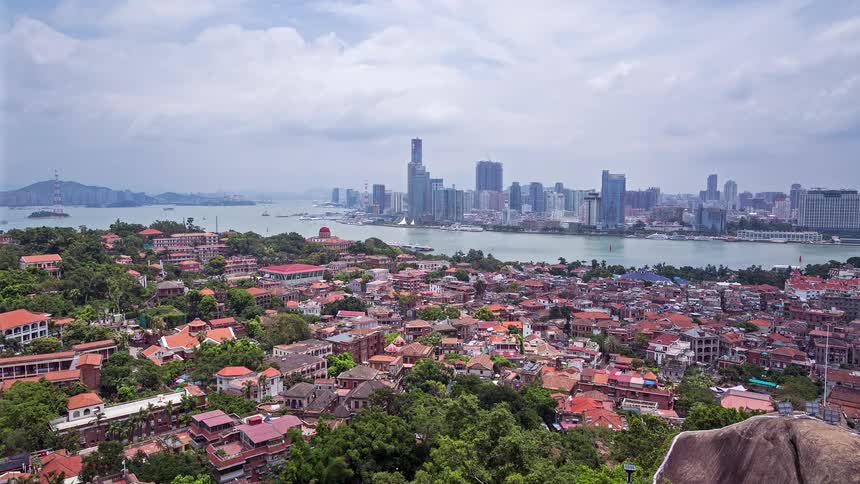 A wide panoramic view shows the red tiled roofs of a coastal city in the foreground with the modern glass skyscrapers rrising across the water under a cloudy sky