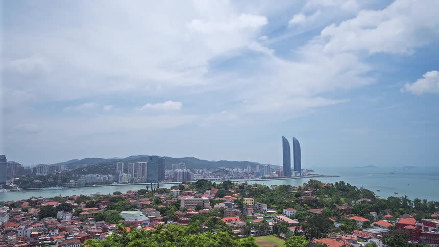 A wide aerial shot shows a coastal city with a mix of traditional red roof houses and modern skyscrapers under a bright cloudy sky while twin towers rise above the urban skyline near the blue ocean water and lush green trees