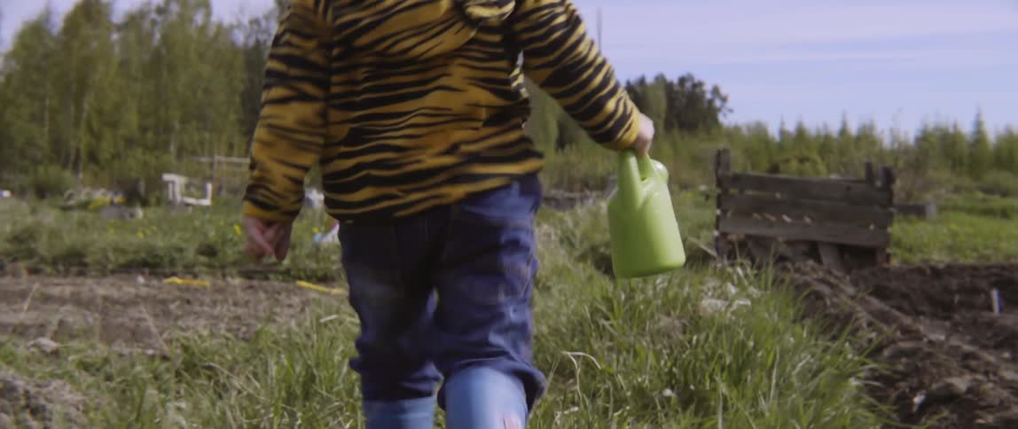 Young child walking through a grassy meadow while holding a small green pitcher.