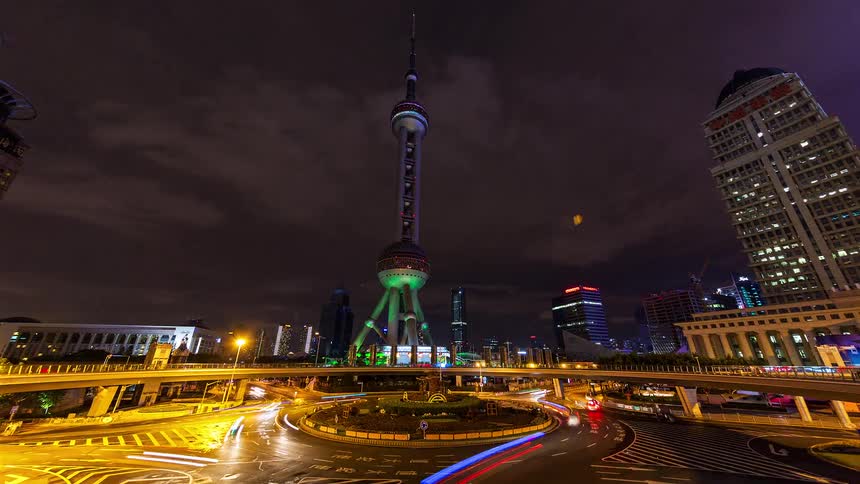 A 360 degree view of the oriental pearl tower and surrounding skyscrapers at night as light trails from traffic circle a busy roundabout under a cloudy sky in the pudong district of shanghai