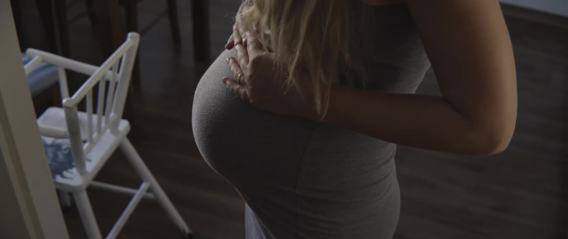 A pregnant woman is feeling her belly as she stands indoors