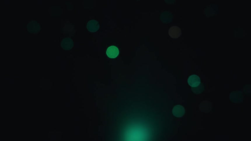 A glowing orb of light shifts through a spectrum of orange green and blue against a black background while soft bokeh circles drift across the frame in a continuous abstract motion of color and light
