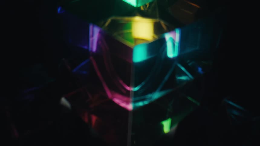 A glass cube rotates slowly on a dark surface while vibrant light refracts through its facets to create colorful rainbows and sharp geometric reflections in a dark room