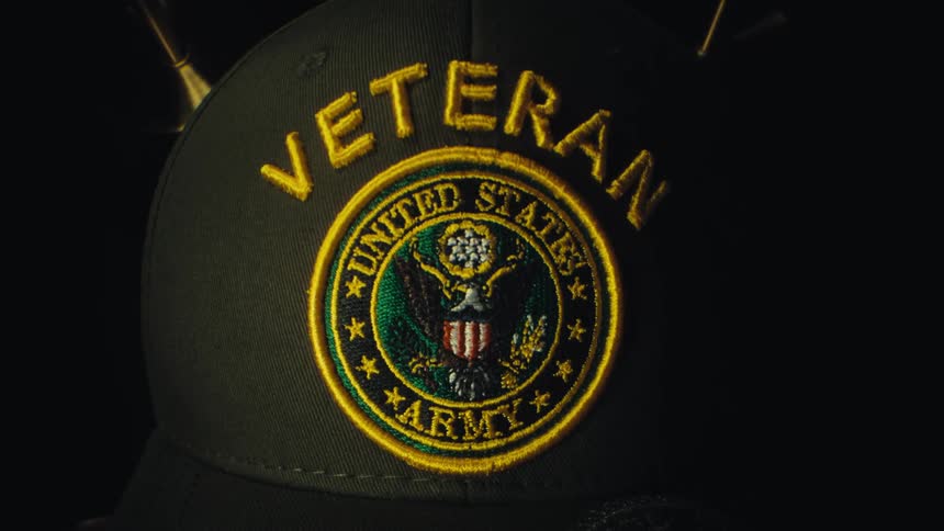 A close view of a dark olive green hat features an embroidered united states army patch with the word veteran stitched in yellow thread above the emblem against a dark background