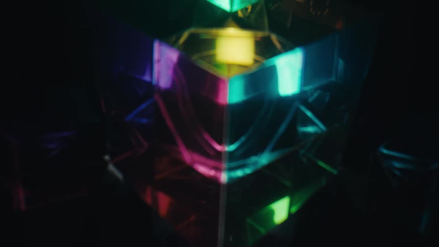 A close view shows a glass prism reflecting vibrant green pink and blue light in a dark space while a bright white beam shines from below