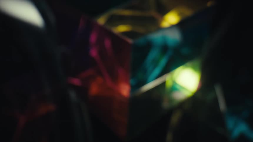 A glass cube prism sits in a dark space as light passes through its facets creating vibrant colorful refractions in blue yellow and magenta