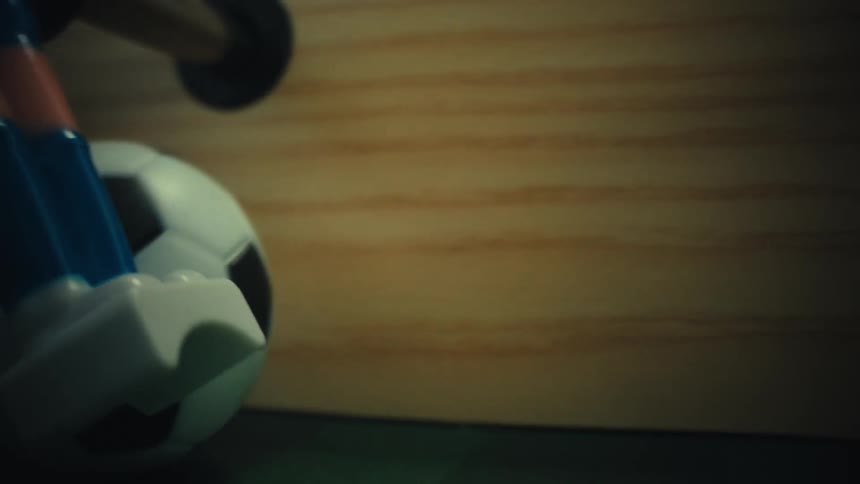 A miniature soccer ball rolls across the green surface of a wooden foosball table while the blue and white feet of a plastic player stay positioned in the blurred foreground