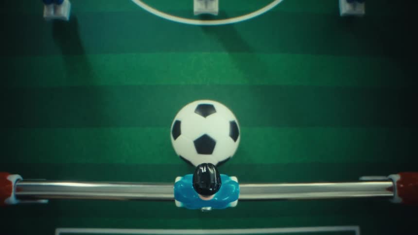 A view from above shows a blue plastic figure on a metal rod hitting a small soccer ball across a green table while playing a game of table football