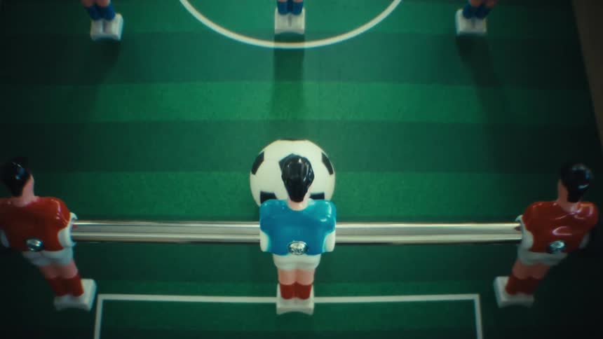 A top down close up shows a black and white soccer ball positioned directly behind a blue foosball player on a green table while red and yellow teammates stand on parallel metal rods across the pitch