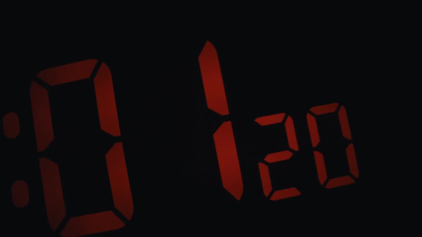 A close up shot shows a digital alarm clock with glowing red led numbers against a pitch black background as the camera moves closer to the illuminated electronic display