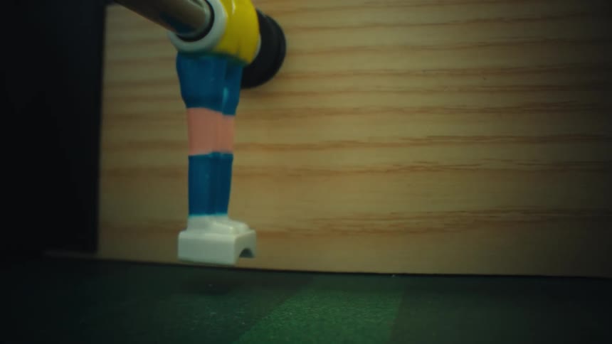 A close view moves past a plastic figure to show a small soccer ball on a green table before ending on a white foot of another player while playing table football