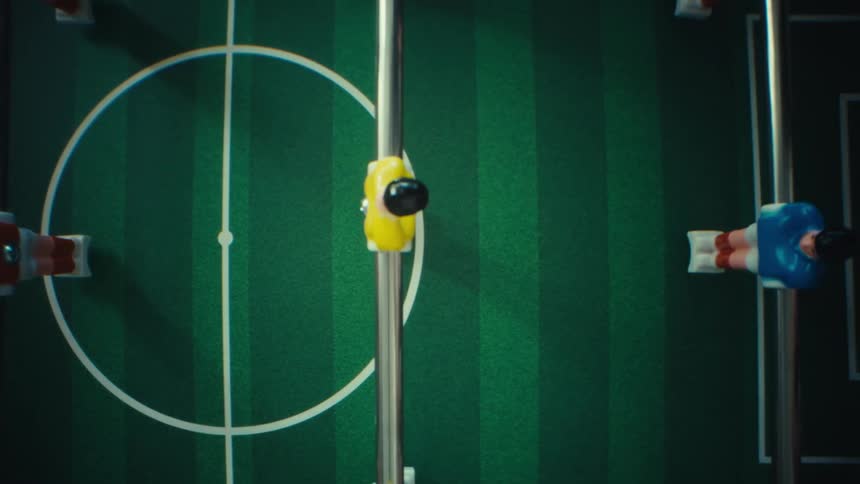 A green foosball table while the rods slide across the field and the miniature players pass over the white center circle and penalty box toward the goal