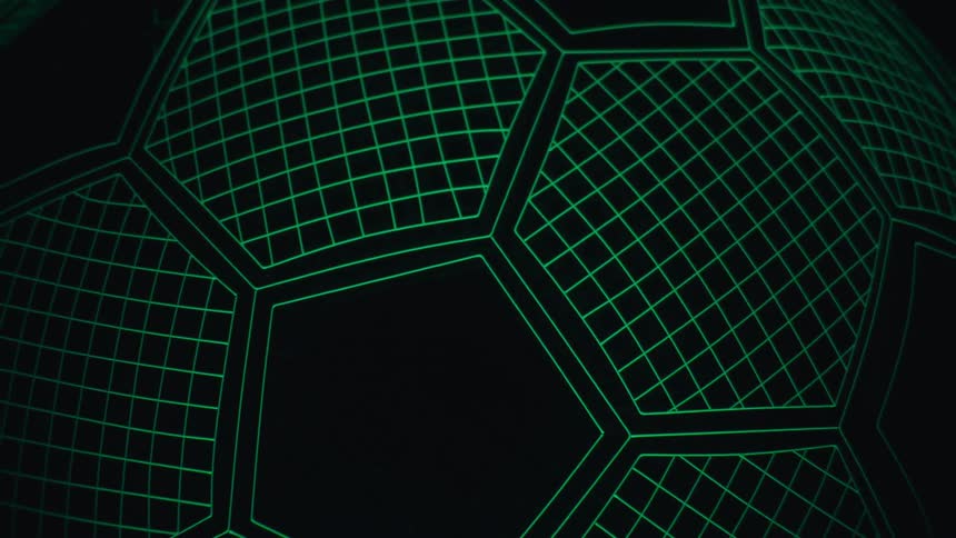 A neon green glowing soccer ball with a grid wireframe pattern slowly zooms out from a close up to reveal its full spherical shape on a flat stand against a dark black background