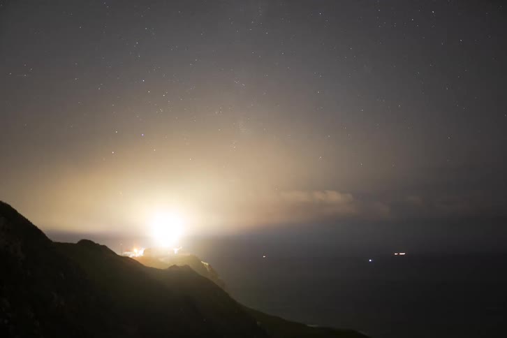 A lighthouse beam rotates across the night sky above a dark coastline with a starry backdrop