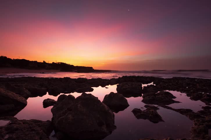 Rocks along the coast as waves roll into shore and the sun glows on the horizon