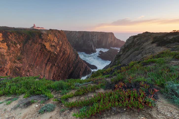 A mountainous hill with a flowing stream beneath it leading into the ocean in the background with short grasses covering the hillside as time moves from sunrise to sunset