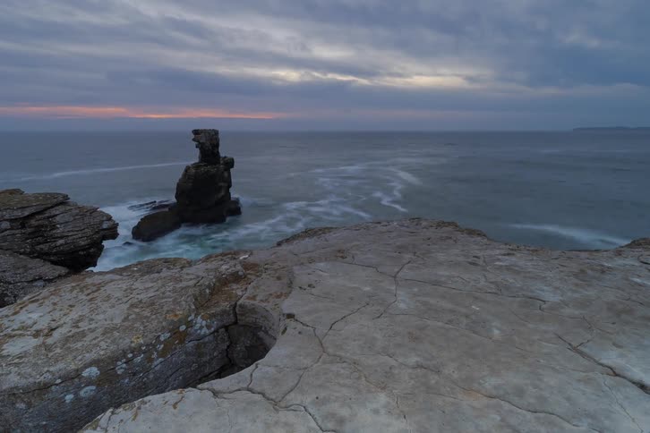 A view from a rocky cliff looks down at waves crashing onto a dark sea with a single large rock formation standing in the water under a cloudy twilight sky