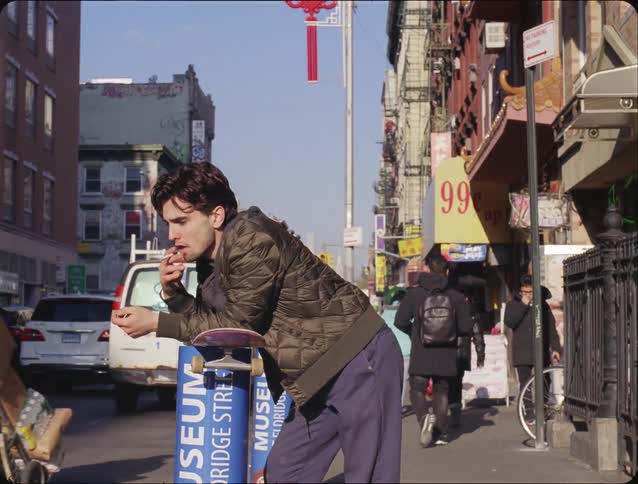 Man leaning on a skateboard smoking a cigarette on a busy city sidewalk