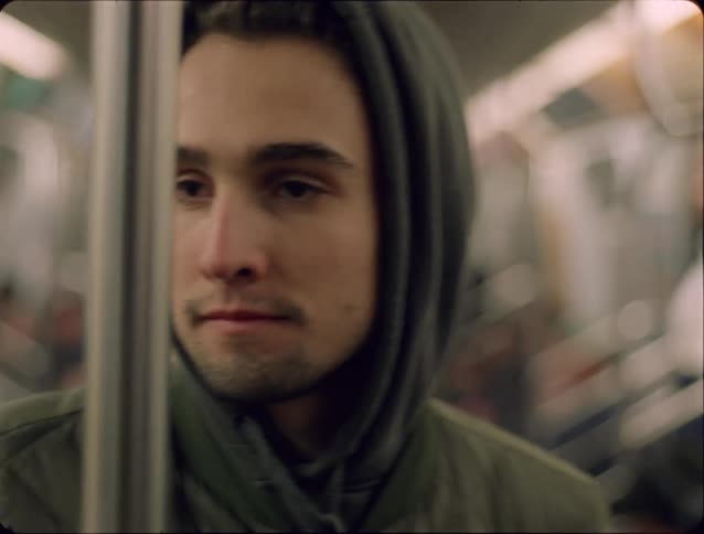 A young man in a hoodie stands quietly on a subway train his tired gaze drifting as the blurred carriage moves around him
