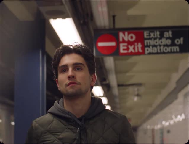 A man with dark hair wearing a quilted jacket and grey hoodie walks along a subway platform under bright lights and a red exit sign in a transit station