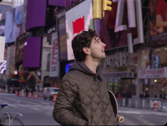 A young man stands in the middle of times square holding a pink skateboard while looking up at the large digital billboards surrounding him