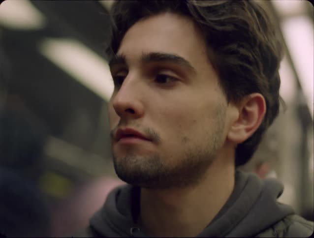 A close view shows a man with dark wavy hair and a beard wearing a grey hoodie while standing on a moving subway train with blurred lights passing by in the background